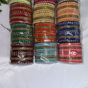 May include: A selection of colourful bangles in shades of green, pink, red, and blue. Each stack is wrapped in clear plastic and features gold-coloured detailing. The bangles are labelled with the number "2.4".