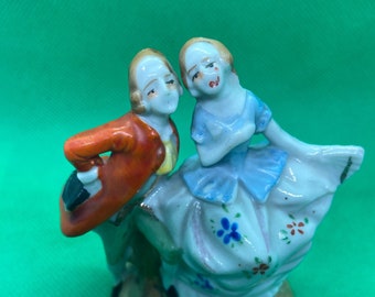 May include: A porcelain figurine of a couple dancing. The woman is wearing a blue and white dress with floral accents, and the man is wearing a red jacket with gold trim. The figurine is hand-painted and has a vintage aesthetic.