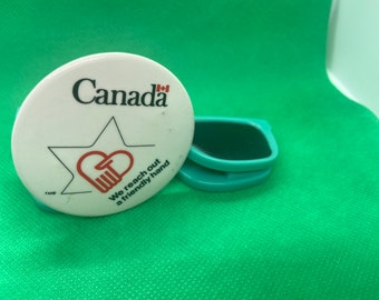 May include: A white round button with the word "Canada" and a red heart with a hand inside. The button also has the text "We reach out a friendly hand" and a small Canadian flag. The button is next to a teal blue plastic clip.