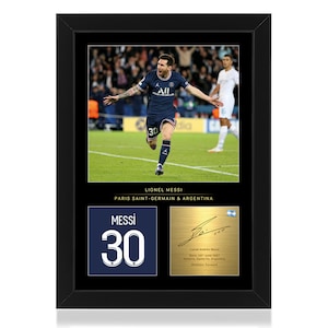 Pode incluir: Uma fotografia emoldurada de Lionel Messi, um jogador de futebol profissional, vestindo uma camisa azul e branca com o número 30. A foto foi tirada em um campo verde com outros jogadores ao fundo. A moldura inclui uma placa dourada com a assinatura de Messi e uma camisa azul com o número 30.
