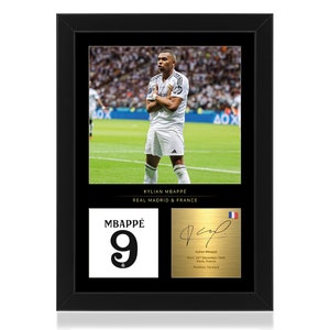 May include: A framed photograph of footballer Kylian Mbappé wearing a white jersey with a black and white number 9 on the front. The photo is set against a green football pitch. The frame is black with a gold trim. The text "KYLIAN MBAPPE REAL MADRID & FRANCE" is printed below the photo. A smaller photograph of Mbappé's signature and jersey number is included in the frame.