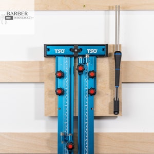 May include: A blue TSO woodworking jig with red knobs and a silver metal rod. The jig is mounted on a wooden wall with a silver metal rod hanging next to it.