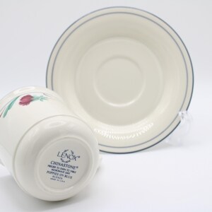 May include: A white Lenox Chinastone teacup and saucer with a blue rim and a floral design. The teacup has the words "Chinastone" and "Poppies on Blue" printed on the bottom.