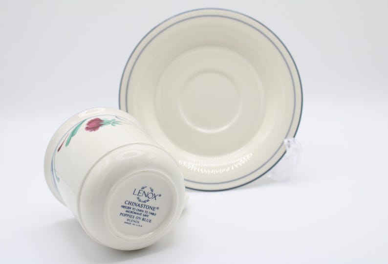 May include: A white Lenox Chinastone teacup and saucer with a blue rim and a floral design. The teacup has the words "Chinastone" and "Poppies on Blue" printed on the bottom.
