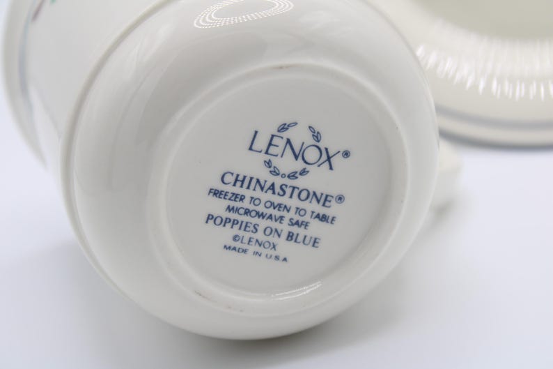 May include: The bottom of a white ceramic mug with blue lettering that reads "LENOX&reg; CHINASTONE&reg; FREEZER TO OVEN TO TABLE MICROWAVE SAFE POPPIES ON BLUE LENOX MADE IN USA"