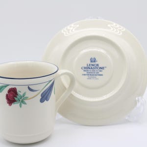 May include: A white teacup and saucer with a floral design in shades of blue, green, and red. The saucer has a raised edge and a scalloped design. The teacup has a blue rim and a floral design on the side. The saucer has the text "LENOX CHINASTONE" in blue on the bottom.