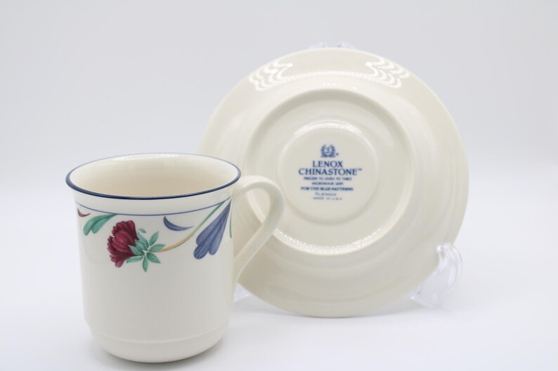 May include: A white teacup and saucer with a floral design in shades of blue, green, and red. The saucer has a raised edge and a scalloped design. The teacup has a blue rim and a floral design on the side. The saucer has the text "LENOX CHINASTONE" in blue on the bottom.