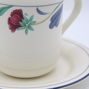 May include: A white ceramic coffee mug with a blue rim and floral design. The mug is sitting on a matching saucer with a blue rim.
