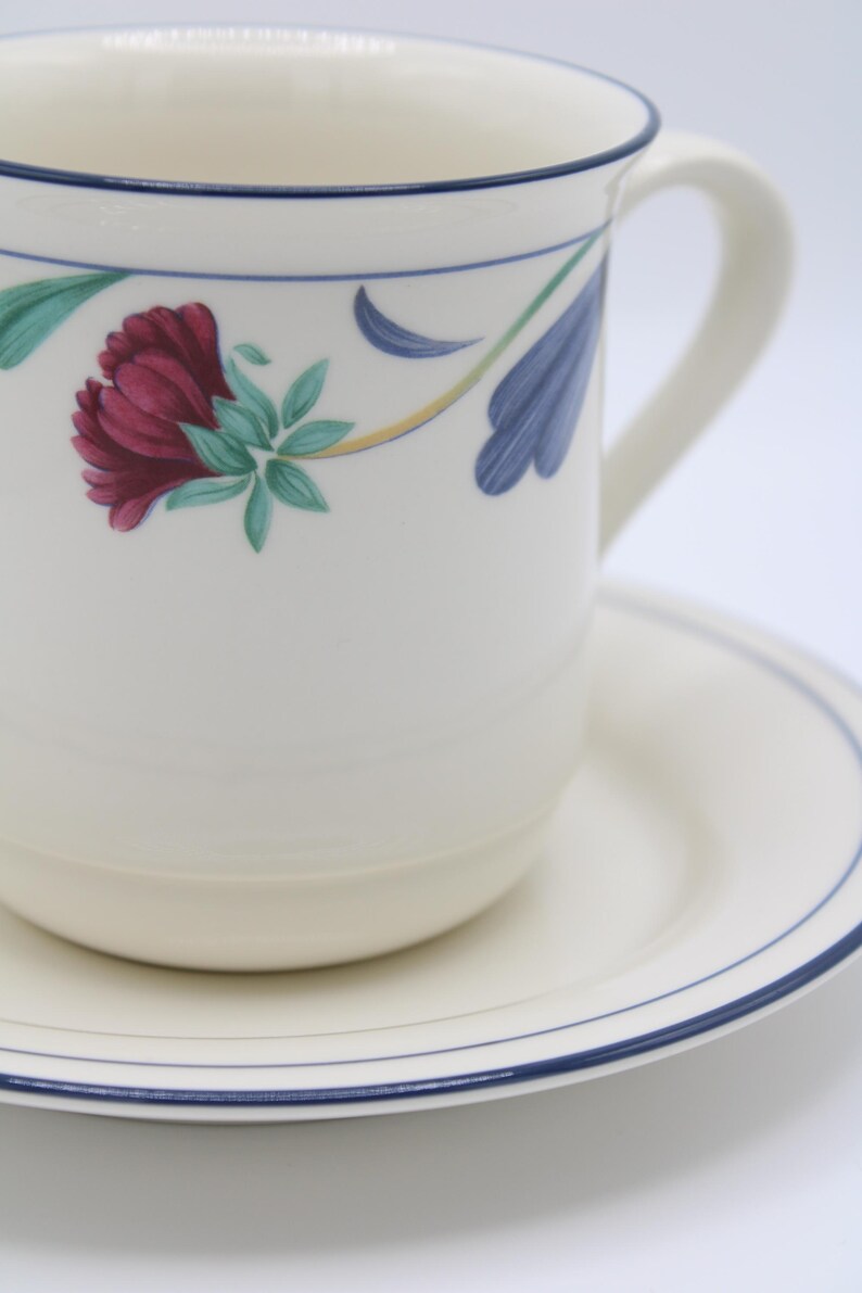 May include: A white ceramic coffee mug with a blue rim and floral design. The mug is sitting on a matching saucer with a blue rim.