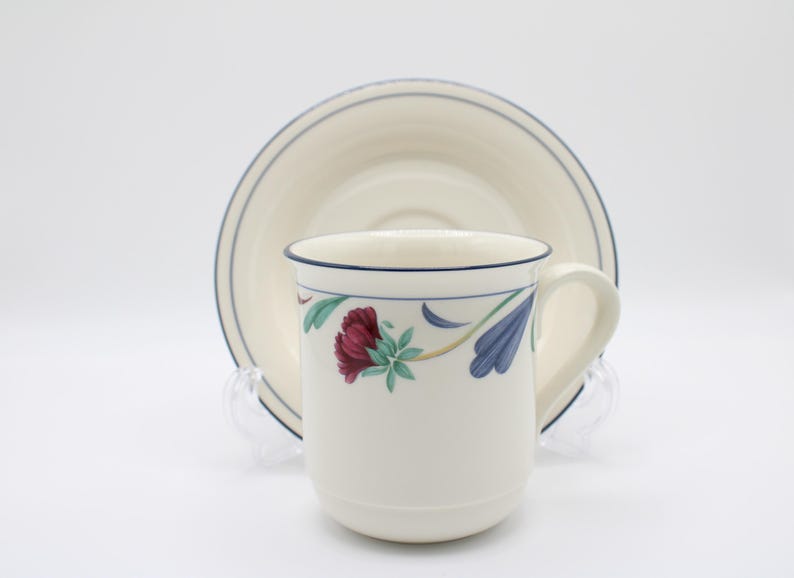 May include: A white ceramic teacup and saucer with a floral design. The cup has a blue rim and a blue and green floral pattern with red flowers. The saucer has a blue rim and a white center.