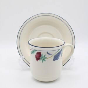 May include: A white ceramic coffee cup with a blue rim and floral design. The cup is sitting on a matching saucer with a blue rim.