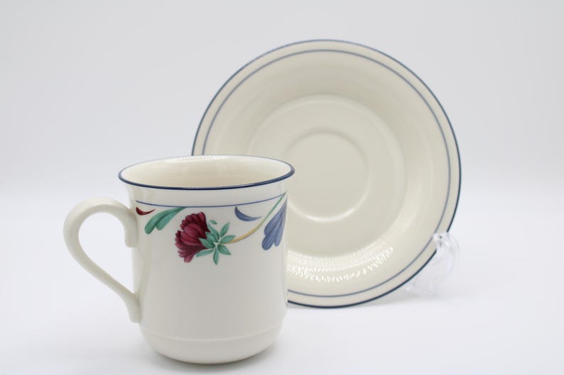 May include: A white ceramic coffee cup and saucer with a floral design. The cup has a blue rim and a blue and green floral pattern. The saucer has a blue rim and a white center.