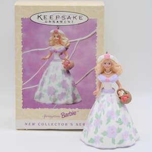 May include: A Springtime Barbie ornament, featuring a doll in a floral dress and a matching hat, holding a basket of flowers. The ornament is displayed next to its original box, which also features the doll.