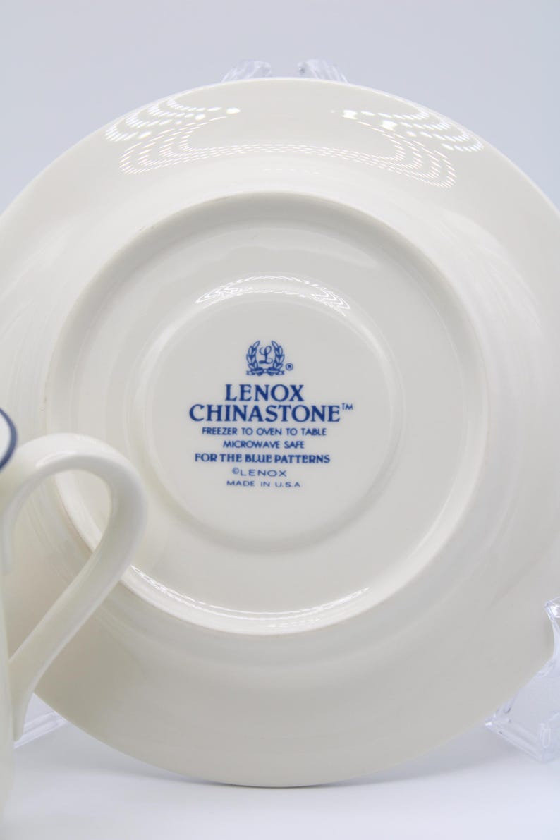 May include: A white saucer with a blue rim and a blue Lenox Chinastone logo. The logo reads "Lenox Chinastone", "Freezer to Oven to Table", "Microwave Safe", "For the Blue Patterns", "Lenox", and "Made in USA".