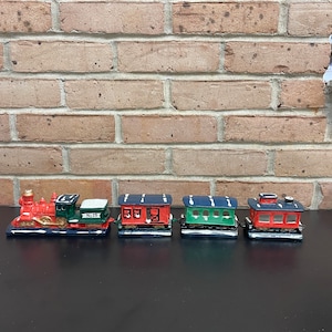 Könnte beinhalten: Ein Miniatur-Weihnachtszug-Set mit einer roten Lokomotive, grünen und roten Waggons und einer blauen Basis. Die Waggons haben weiße Akzente und sind auf einer schwarzen Oberfläche angeordnet. Der Hintergrund ist eine Backsteinmauer.
