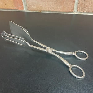 Puede incluir: Pinzas de servir plateadas con diseño de tijera. Un extremo es plano y en forma de espátula, el otro en forma de tenedor. Están unidos por un pivote central. Los mangos tienen orificios circulares para los dedos. Fondo negro y pared de ladrillo.