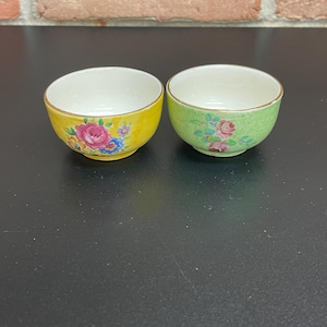 May include: Two small porcelain teacups with floral designs. One cup is yellow with a pink rose and other flowers. The other cup is green with a pink rose and other flowers.