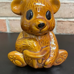 Puede incluir: Un tarro de miel de cerámica marrón con forma de oso, con ojos negros y una expresión sonriente. El oso sostiene un tarro de miel con la palabra "HONEY" en relieve. El tarro está sobre una superficie negra.