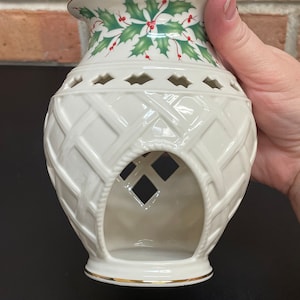 May include: Cream-colored ceramic candle holder with a woven basket design. The top features a green holly and red berry pattern with a gold rim. The holder has diamond-shaped cutouts and a large opening in the front, perfect for a festive holiday display.