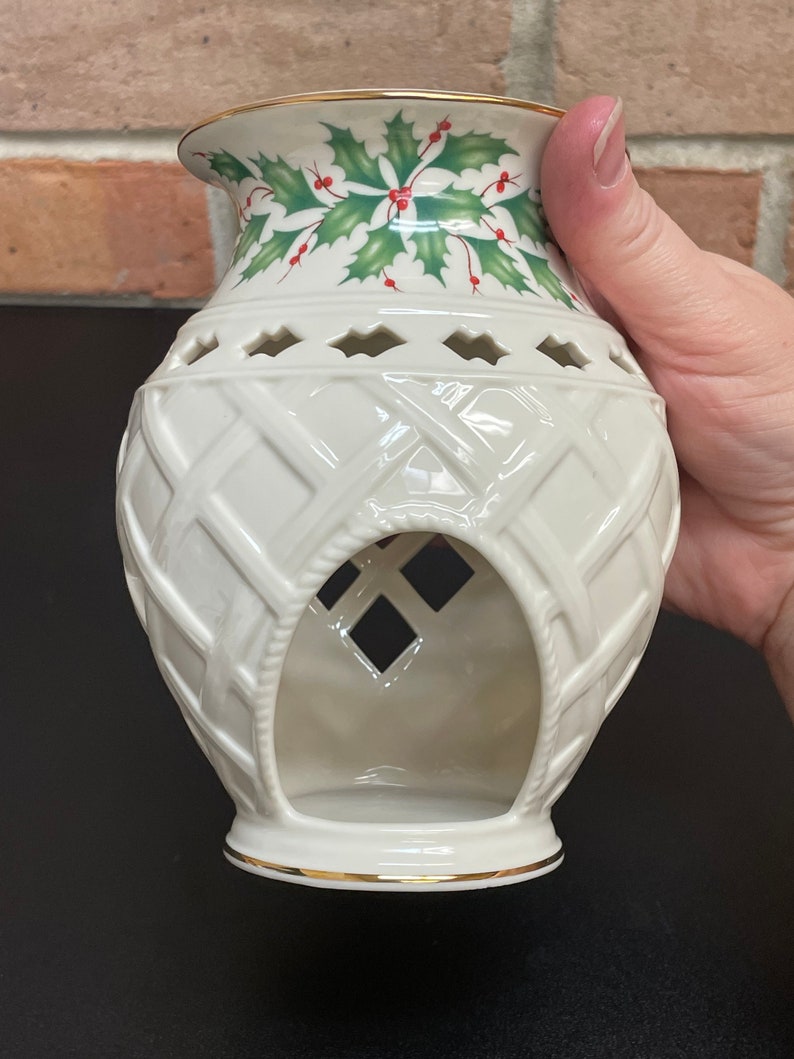 May include: Cream-colored ceramic candle holder with a woven basket design. The top features a green holly and red berry pattern with a gold rim. The holder has diamond-shaped cutouts and a large opening in the front, perfect for a festive holiday display.