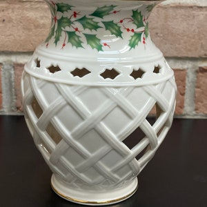 May include: Off-white ceramic vase with a lattice design and gold trim. The top of the vase features a holly and berry pattern in green and red. The vase has a flared top and a round base.