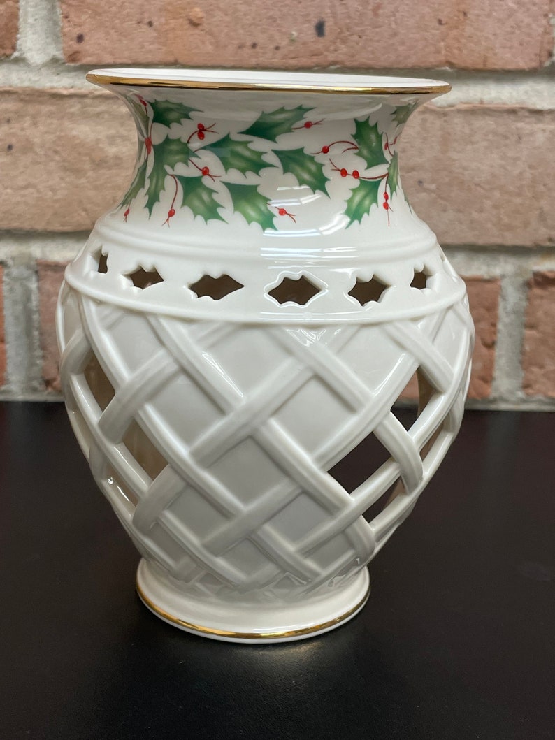 May include: Off-white ceramic vase with a lattice design and gold trim. The top of the vase features a holly and berry pattern in green and red. The vase has a flared top and a round base.