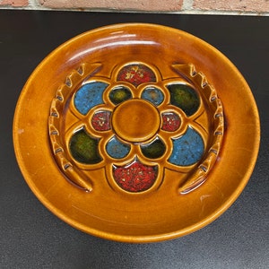 May include: A brown ceramic ashtray with a multi-colored floral design. The ashtray has a central circular depression and four smaller depressions around the edge.