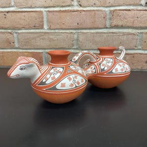 May include: Two terracotta-colored ceramic vessels with intricate white, black, and orange patterns. One vessel has a handle and a spout, while the other has a stylized animal head. The pottery is likely handmade.