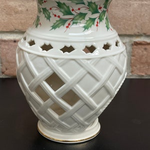 May include: A white ceramic vase with a lattice design and a gold rim. The top of the vase features a holly and berry pattern in green and red. The vase is a decorative item.