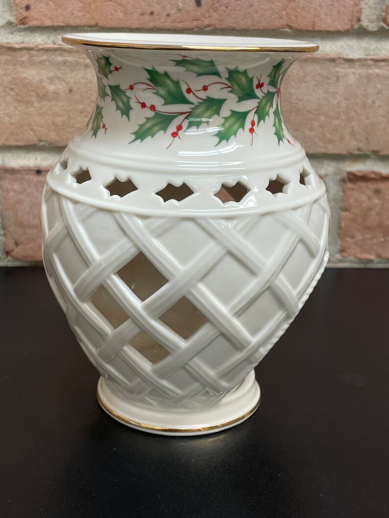 May include: A white ceramic vase with a lattice design and a gold rim. The top of the vase features a holly and berry pattern in green and red. The vase is a decorative item.