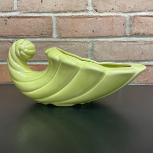 May include: A light green ceramic planter with a unique, curved design. The planter has a spiral detail on one end and a long, open trough for planting. The smooth, glossy finish adds to its decorative appeal. This piece is suitable for indoor or outdoor use.