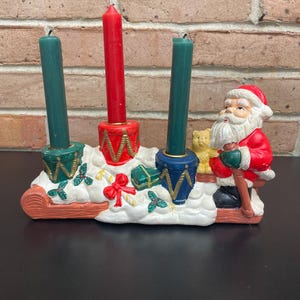 Puede incluir: Un portavelas navideño con Papá Noel, tres velas y elementos decorativos. Las velas son rojas y verdes, y el soporte tiene un diseño festivo con un lazo rojo y regalos verdes envueltos. Papá Noel lleva un traje y un gorro rojos.
