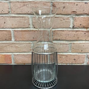 Könnte beinhalten: Eine klare Glasvase mit einem silbernen Drahtkäfig drumherum. Die Vase steht auf einer schwarzen Oberfläche.