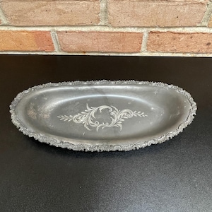 Könnte beinhalten: Ovale, silberfarbene Metallschale mit verzierten, gewellten Kanten. Die Schale hat ein geätztes Blumendesign in der Mitte. Sie wird auf einer schwarzen Oberfläche mit einer Backsteinmauer im Hintergrund präsentiert.