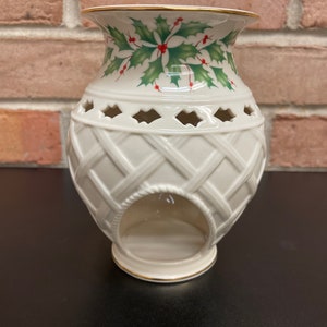 May include: Cream-colored ceramic candle holder with a woven basket design. The top features a holly and berry pattern with green leaves and red berries. The rim and base are trimmed with gold. A decorative piece for the home.