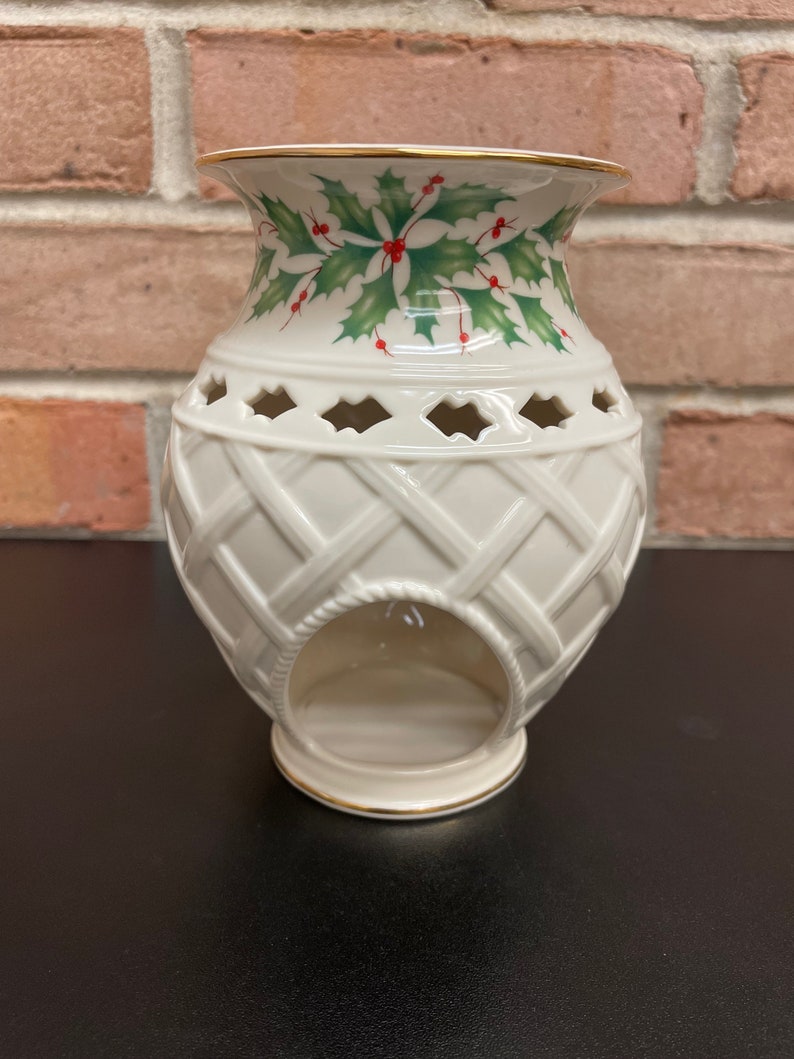 May include: Cream-colored ceramic candle holder with a woven basket design. The top features a holly and berry pattern with green leaves and red berries. The rim and base are trimmed with gold. A decorative piece for the home.