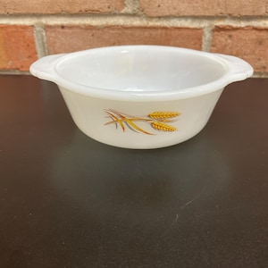 Könnte beinhalten: Weißes, vintage Pyrex-Schüssel mit zwei Griffen und einem Weizen-Design in Gelb und Gold. Die Schüssel ist leer und steht auf einer dunklen Oberfläche vor einer Backsteinmauer. Ein klassisches Küchenutensil.