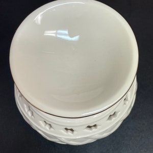 May include: Cream-colored ceramic oil warmer with a round top and a decorative base. The base features a diamond pattern with star-shaped cutouts and a gold trim. The warmer sits on a black surface.