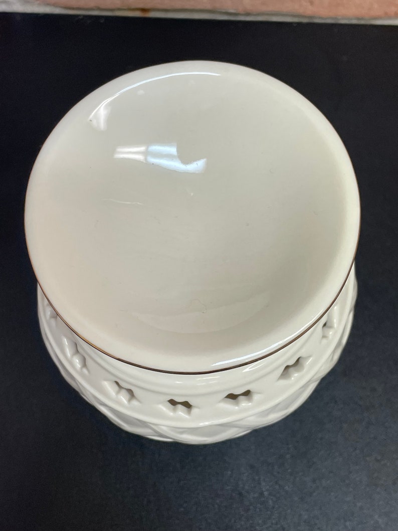 May include: Cream-colored ceramic oil warmer with a round top and a decorative base. The base features a diamond pattern with star-shaped cutouts and a gold trim. The warmer sits on a black surface.