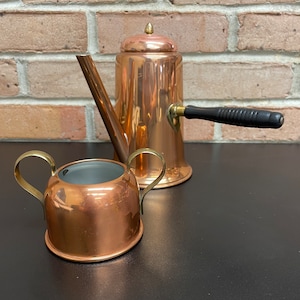 May include: A copper coffee pot with a black handle and a matching copper creamer. The coffee pot has a lid with a small knob on top. The creamer has two handles.