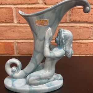 Op de afbeelding: Een vintage, lichtblauwe keramische vaas met een zeemeerminfiguur. De vaas heeft een hoornachtige vorm met een gekruld uiteinde. De zeemeermin knielt en ondersteunt de vaas. De vaas heeft een gouden label met "Royal Haeger".