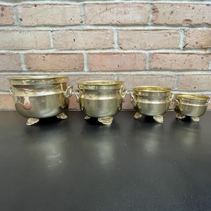 May include: Set of four gold-tone metal planters with feet. The planters are arranged in order of size, with the largest planter on the left and the smallest planter on the right.
