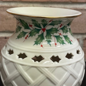 May include: Cream-colored ceramic vase with a gold rim and a decorative holly and berry design. The vase features a lattice pattern and cut-out shapes. A festive holiday home decor item.