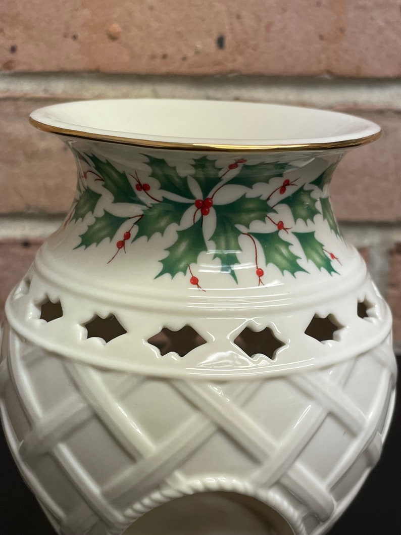 May include: Cream-colored ceramic vase with a gold rim and a decorative holly and berry design. The vase features a lattice pattern and cut-out shapes. A festive holiday home decor item.