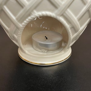 May include: Cream-colored ceramic candle warmer with a basket weave design. The warmer has a round opening to hold a tea light candle. A gold trim accents the base. The candle is unlit.