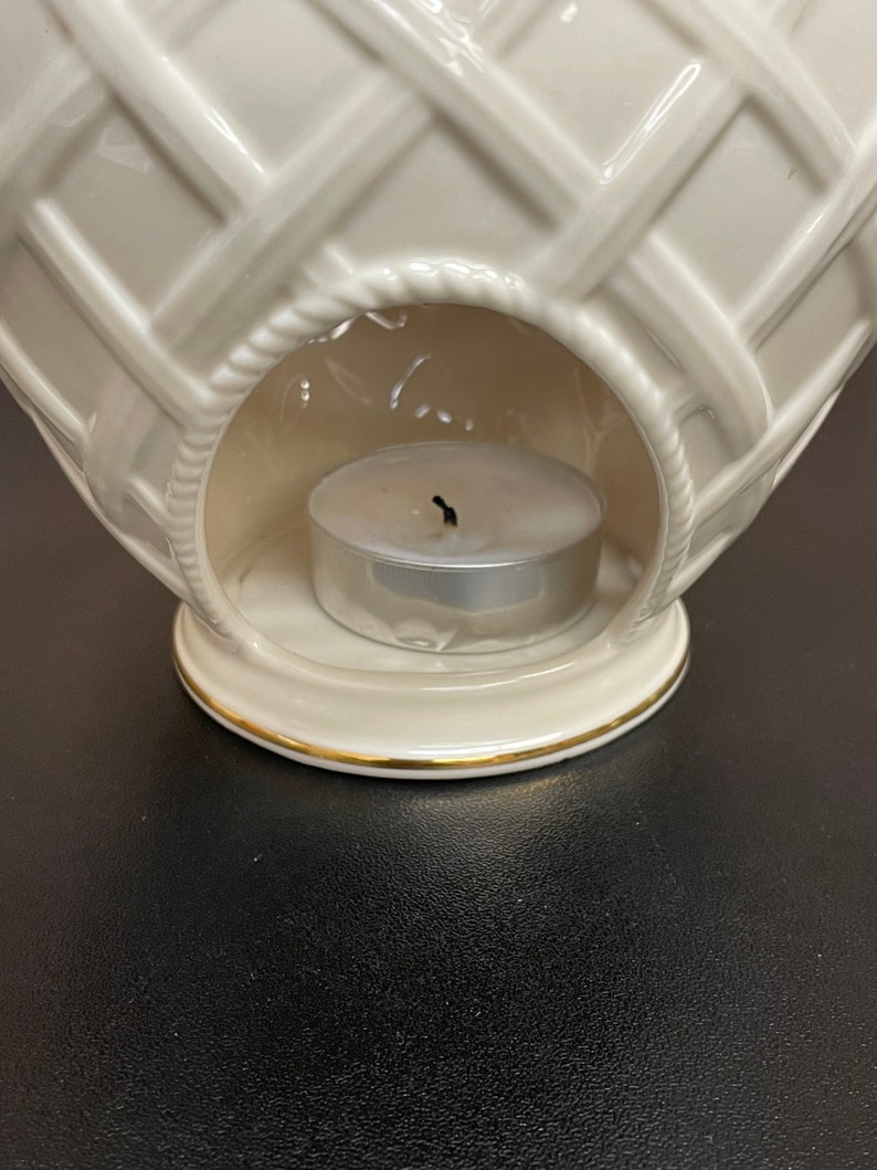 May include: Cream-colored ceramic candle warmer with a basket weave design. The warmer has a round opening to hold a tea light candle. A gold trim accents the base. The candle is unlit.