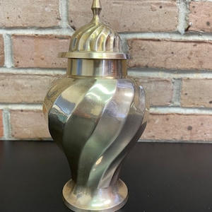 Peut inclure: Un pot décoratif en métal doré avec un motif en spirale et un couvercle. Le pot a une base ronde et un sommet pointu.