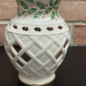 May include: Cream-colored ceramic vase with a lattice design and holly leaf and berry pattern around the top. The rim and base are trimmed with gold. The vase has a decorative, openwork design.