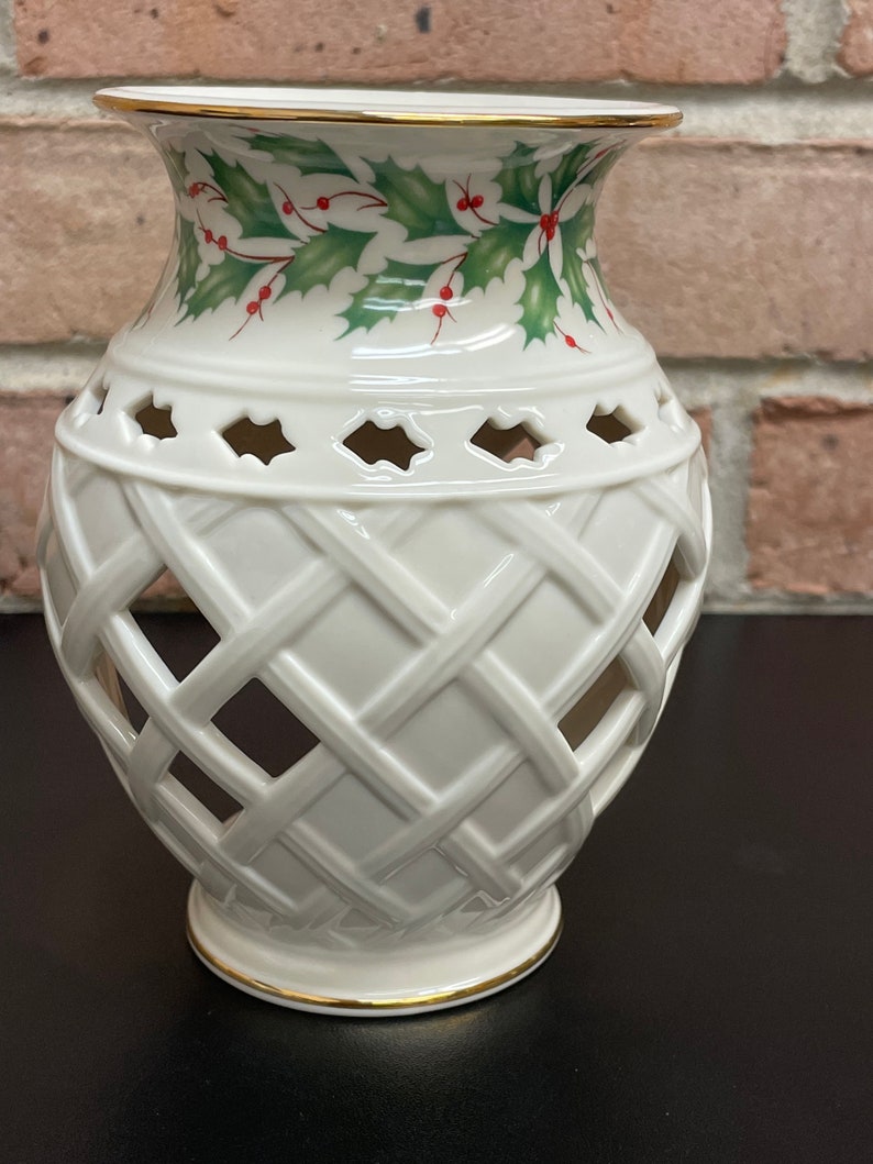 May include: Cream-colored ceramic vase with a lattice design and holly leaf and berry pattern around the top. The rim and base are trimmed with gold. The vase has a decorative, openwork design.