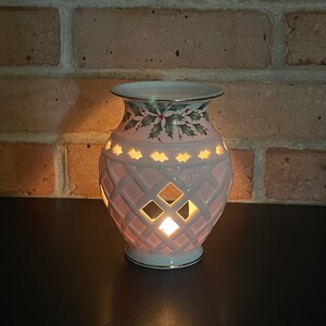 May include: A lit, off-white ceramic candle holder with a diamond lattice pattern. The top rim features a holly and berry design. The candle holder is on a dark surface, with a brick wall in the background.